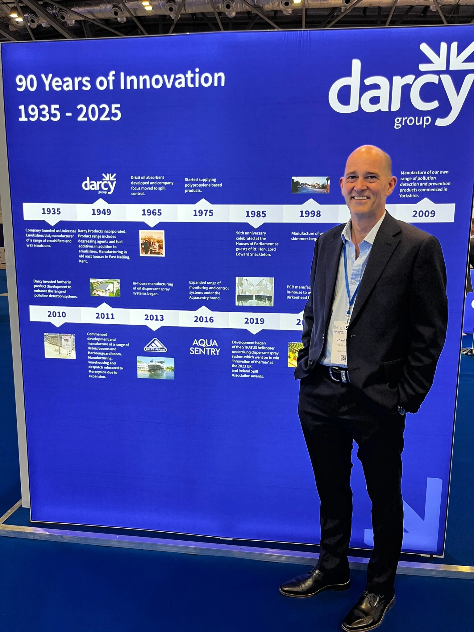Group managing director of the Darcy Group, Richard Proctor, stands in front of a lightbox exhibition stand at Interspill 2025. The lightbox graphic has the title, 90 Years of Innovation 1935 - 2025, and shows a timeline of Darcy's history.